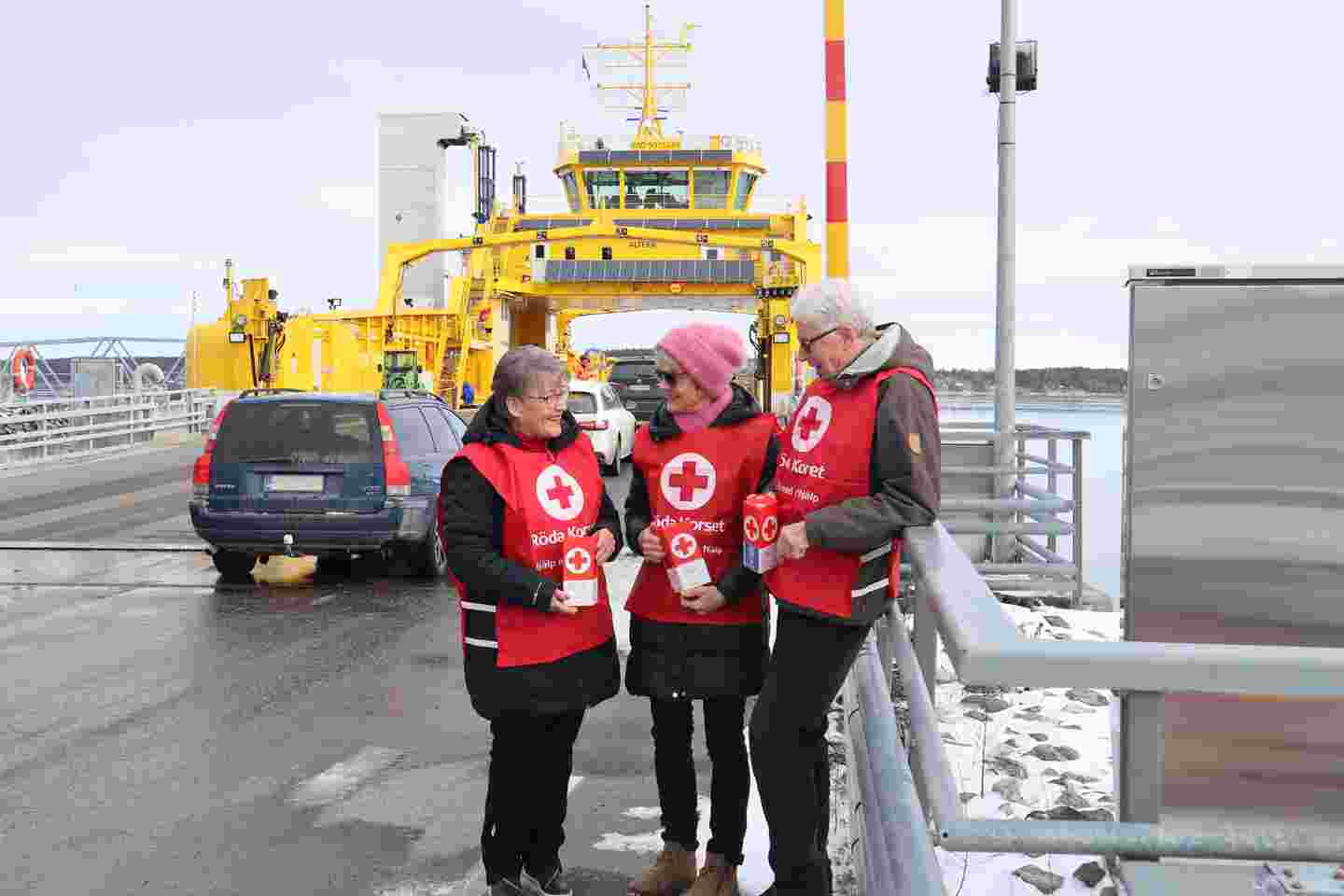 Tre frivilliga i insamlingsvästar och med röda bössor. Det är vinter och i bakgrunden en gul färja