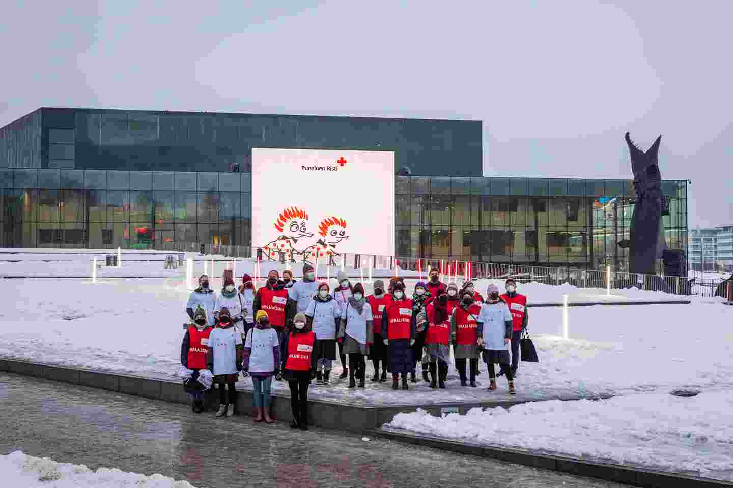 Ryhmäkuva, jossa Punaisen Ristin vapaaehtoiset ja kumpppanit poseeraavat Musiikkitalon valotaululla olevan Punaisen Ristin ja Moomin by Arabian yhteistyökampanjaan liittyvän kuvan edessä.