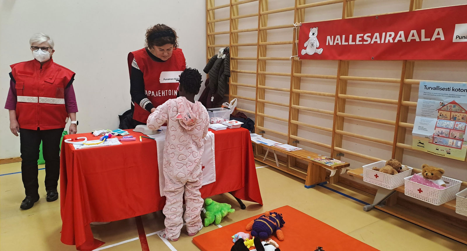 Kaksi henkilöä pukeutuneena Punaisen Ristin liiveihin opastaa Nallesairaalassa lapselle ensiapua.