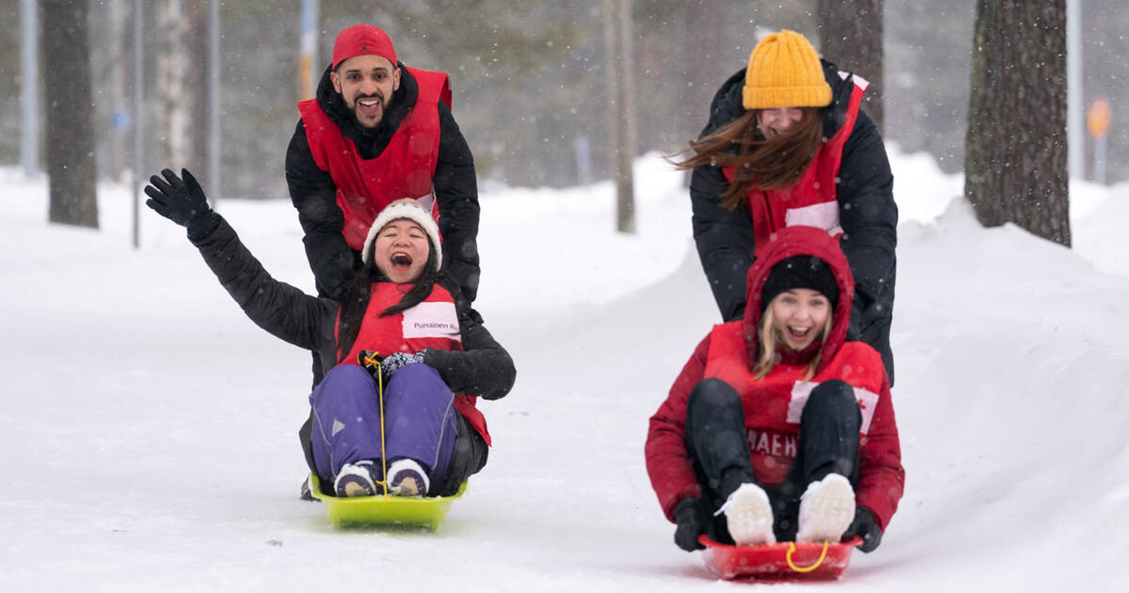 Punaisen Ristin liiveihin pukeutuneet nuoret laskevat pulkalla talvisessa maisemassa.  