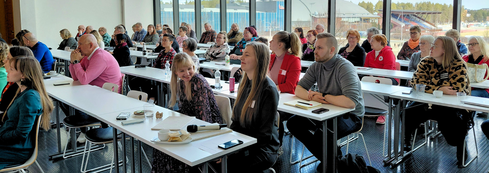 Vapaaehtoiset pohtivat järjestön asioita yhdessä. Valokuvassa vapaaehtoisia ja työntekijöitä pitkien pöytien ääressä, etualalla naurava nainen, taustalla isot ikkunat.