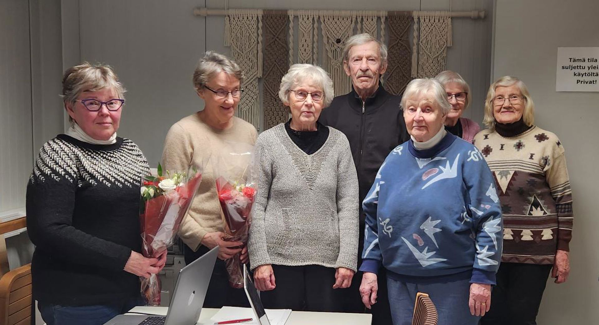 Viisi henkilöä seisoo rivissä sisätiloissa. Kahdella henkilöllä on kädessää kukkakimput.