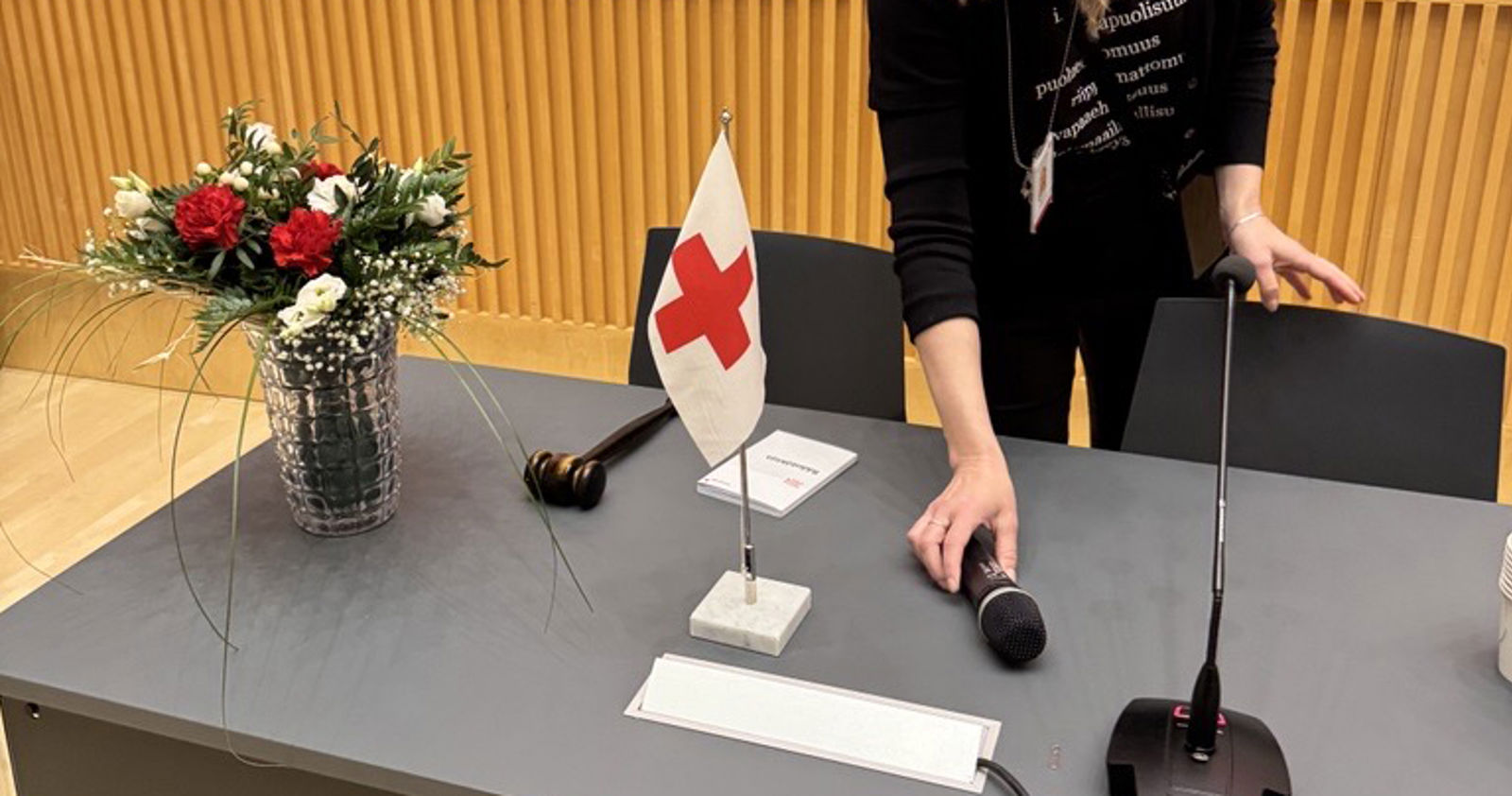 Henkilö seisoo pöydän ääressä, jossa Punaisen Ristin viiri, kukkia ja mikrofoni.