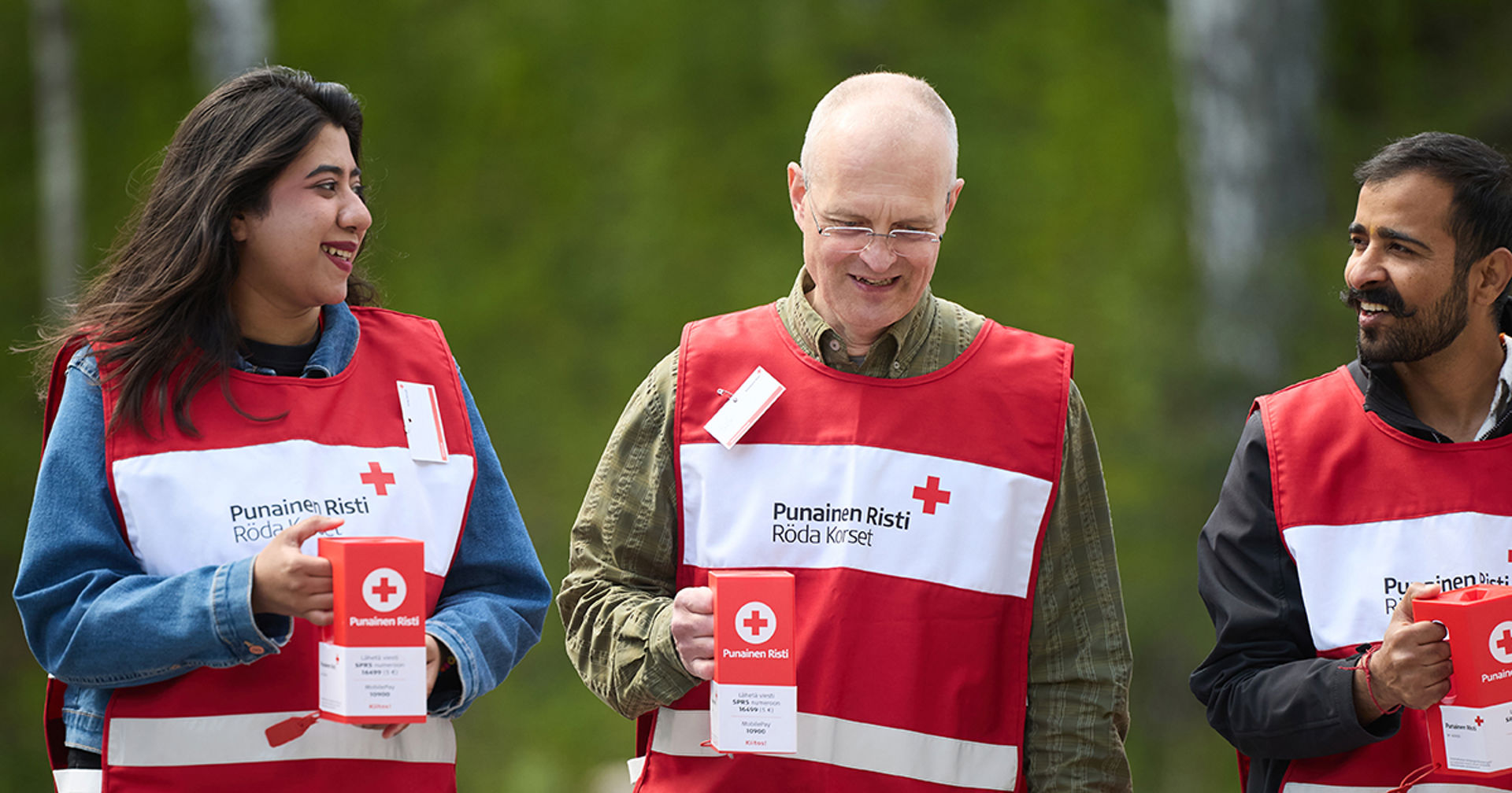 Kolme hymyilevää Punaisen Ristin liiveihin pukeutunutta henkilöä ulkona Punaisen Ristin keräyslippaat käsissään.