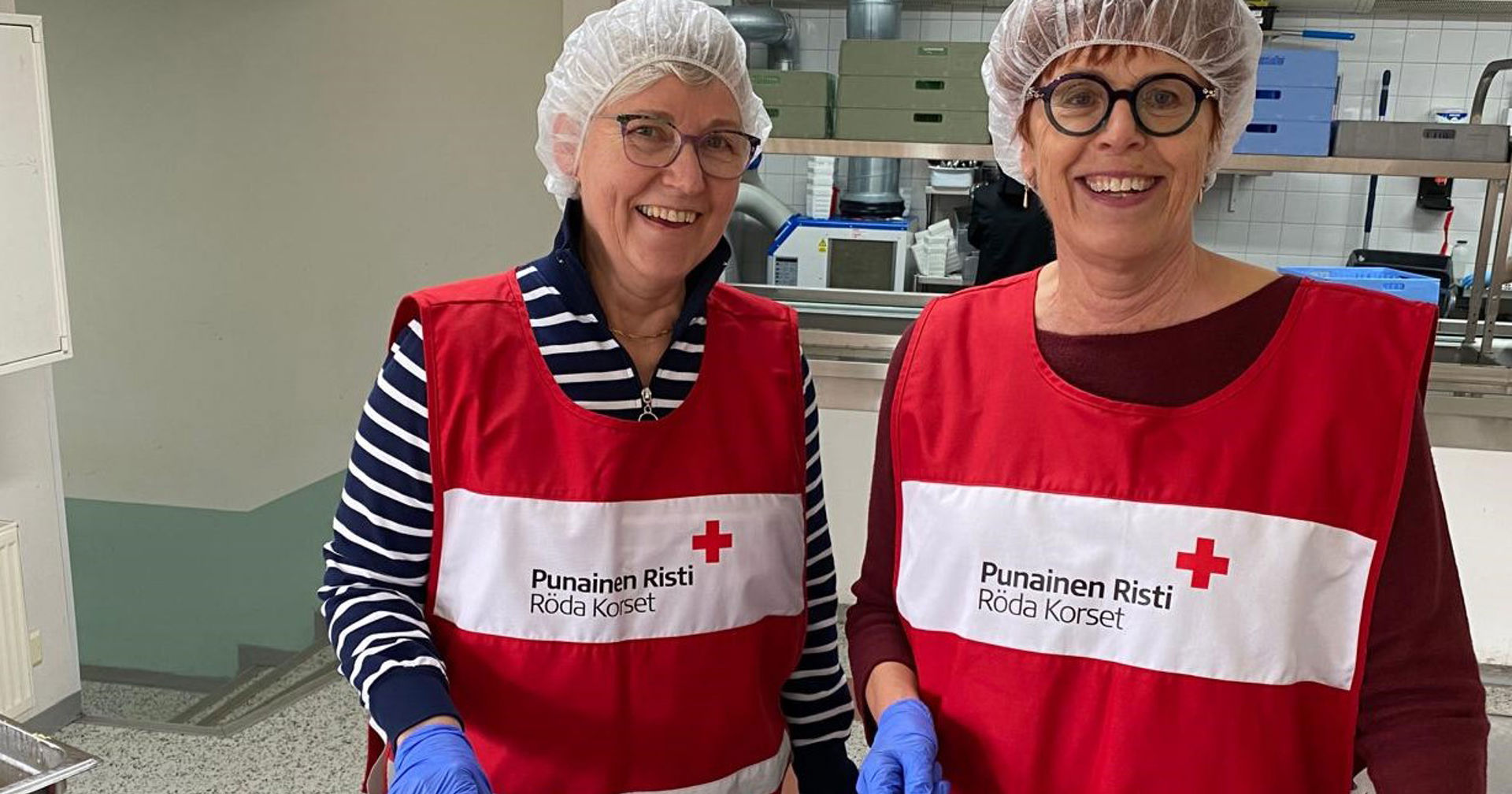 Kaksi hymyilevää Punaisen Ristin vapaaehtoista koulun ruokalassa. 
