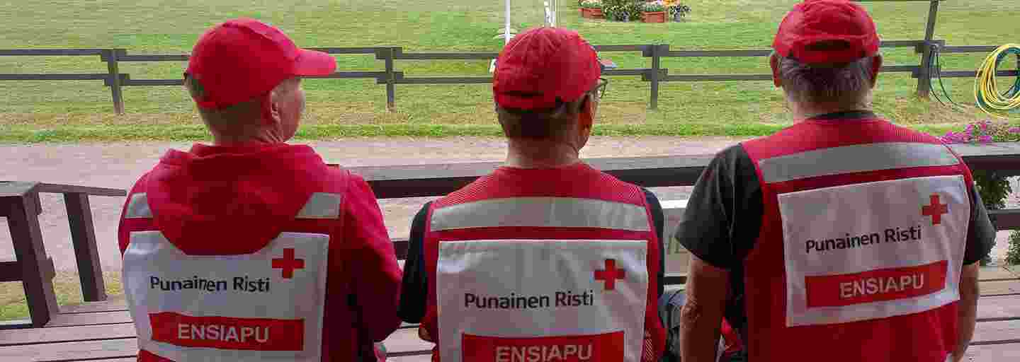 Punaisen Ristin kolme ensiapupäivystäjää istuu ulkona katsomossa selkä kohti kameraa katsoen vihreälle kentälle. Selässä lukee Punainen Risti, Ensiapu.