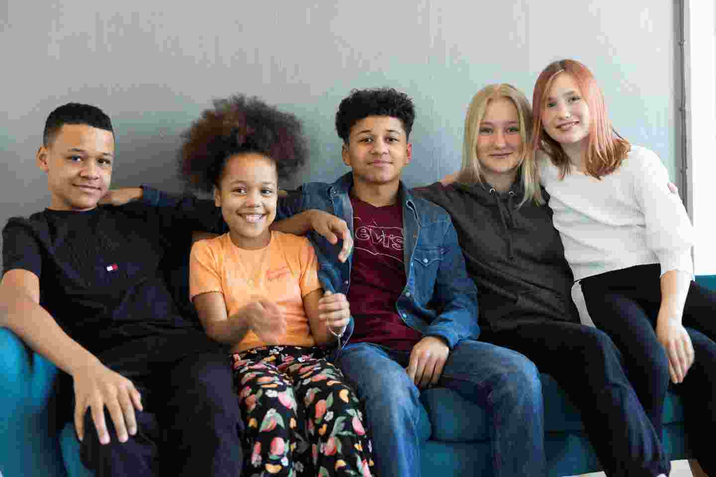 Viisi nuorta istuu sohvalla vierekkäin ja hymyilevät