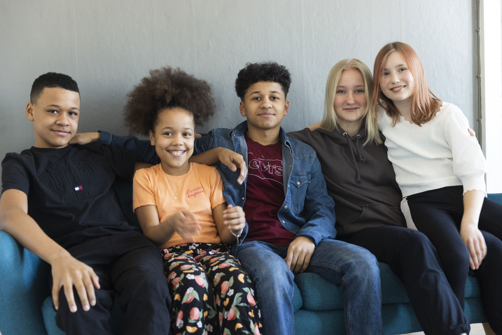 Viisi nuorta istuu sohvalla vierekkäin ja hymyilevät