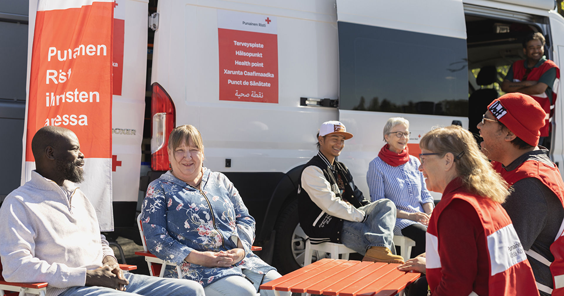 Hymyileviä henkilöitä istumassa tuoleilla ja pöydän ääressä retkeilyauton edessä aurinkoisessa säässä.