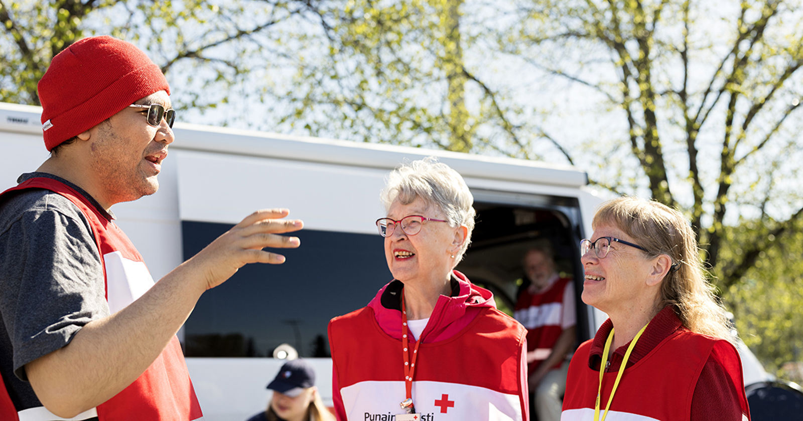 Punaisen Ristin vapaaehtoisia juttelemassa ulkona kevätsäässä. Taustalla valkoinen retkeilyauto.